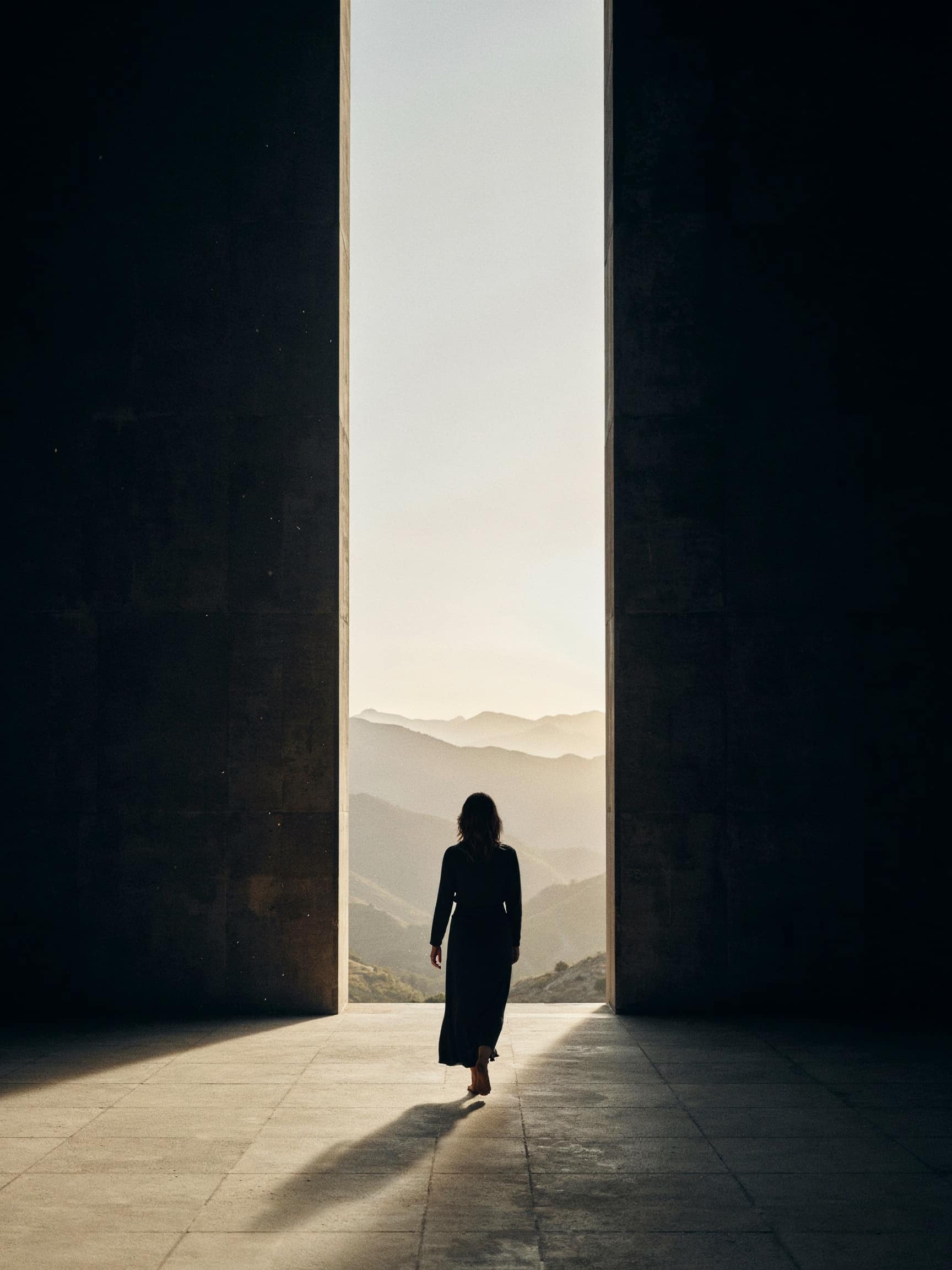 A doorway of dawn between two walls of dark stone; a woman seen from behind walks into the light toward distant mountains.